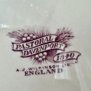 May include: A cream-coloured ceramic surface featuring a dark purple printed maker's mark. The design includes a ribbon banner with the text PASTORAL DAVENPORT 1840, surrounded by wheat stalks and berries, with A.J. WILKINSON LTD, ENGLAND printed below.