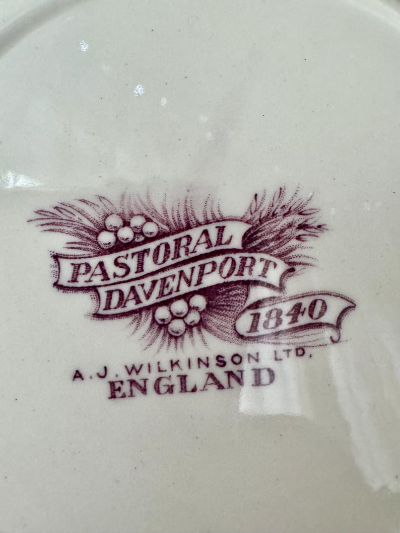 May include: A cream-coloured ceramic surface featuring a dark purple printed maker's mark. The design includes a ribbon banner with the text PASTORAL DAVENPORT 1840, surrounded by wheat stalks and berries, with A.J. WILKINSON LTD, ENGLAND printed below.