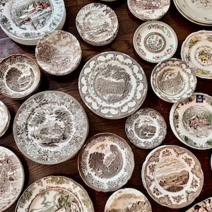 May include: A collection of assorted ceramic plates and bowls featuring various transferware patterns in brown, green, and multi-colored designs. The pieces display detailed landscape scenes, architectural structures, and floral motifs with scalloped edges.