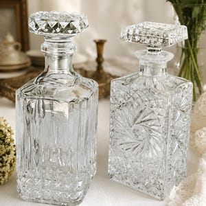 May include: Two clear glass decanters with intricate diamond and geometric patterns. One has a round stopper, the other a square stopper. The decanters are on a white surface, with flowers in the background.