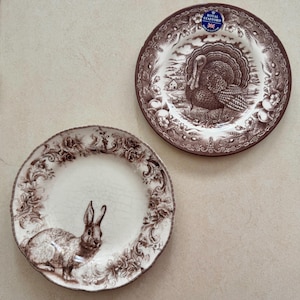 May include: Two round ceramic plates with brown transferware patterns. One plate features a turkey in a rural landscape with a Royal Stafford sticker. The other plate displays a rabbit surrounded by a detailed floral and scroll border.