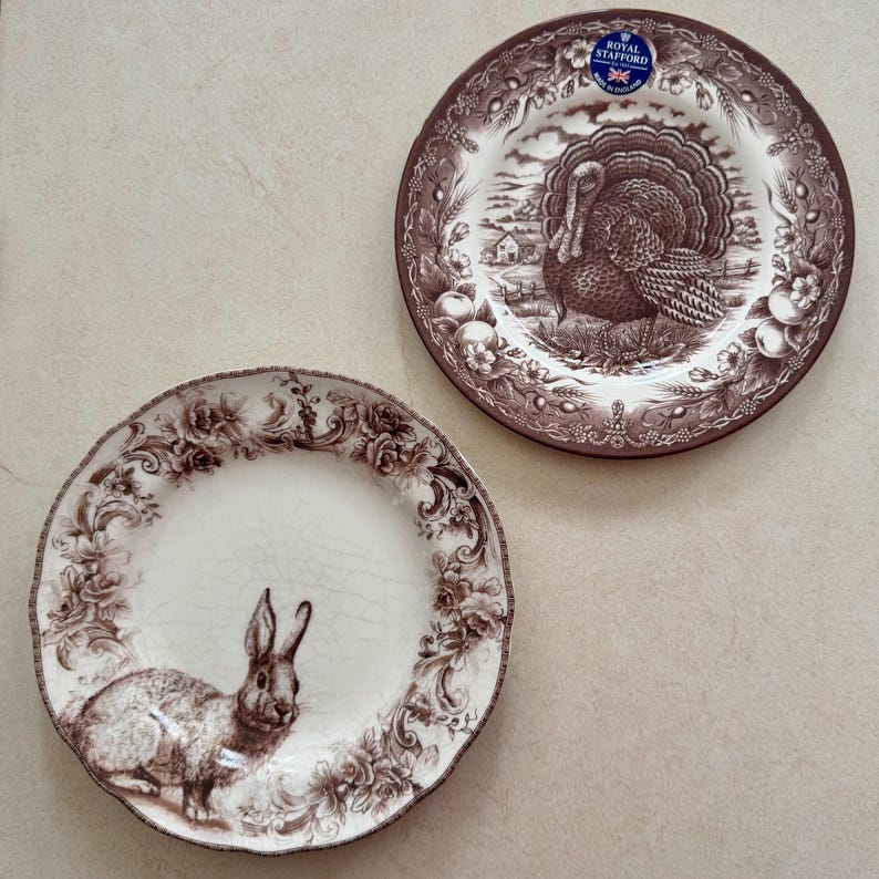 May include: Two round ceramic plates with brown transferware patterns. One plate features a turkey in a rural landscape with a Royal Stafford sticker. The other plate displays a rabbit surrounded by a detailed floral and scroll border.