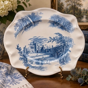 May include: An oval ceramic serving platter with a scalloped edge, featuring a white base and blue transferware illustrations of a castle, horse-drawn carriage, and garden scenes. The platter is displayed on a gold-toned metal stand.
