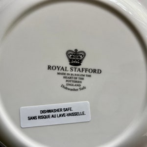 K&ouml;nnte beinhalten: Ein wei&szlig;er Keramikteller mit einem schwarz-wei&szlig;en Logo f&uuml;r Royal Stafford, hergestellt in Burslem, England. Das Logo zeigt eine Krone und den Text "Royal Stafford" und "Dishwasher Safe". Ein wei&szlig;er Aufkleber auf der Unterseite des Tellers lautet "Dishwasher Safe. Sans Risque Au Lave-Vaissele."