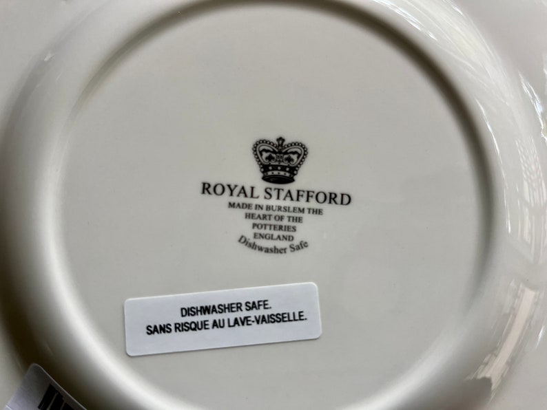 K&ouml;nnte beinhalten: Ein wei&szlig;er Keramikteller mit einem schwarz-wei&szlig;en Logo f&uuml;r Royal Stafford, hergestellt in Burslem, England. Das Logo zeigt eine Krone und den Text "Royal Stafford" und "Dishwasher Safe". Ein wei&szlig;er Aufkleber auf der Unterseite des Tellers lautet "Dishwasher Safe. Sans Risque Au Lave-Vaissele."