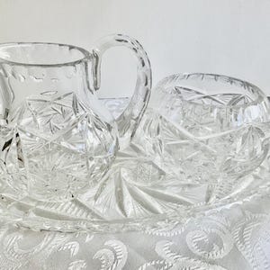 May include: A clear cut glass creamer pitcher and a matching sugar bowl set resting on an oval glass tray. Each piece features a detailed starburst and geometric faceted pattern with scalloped edges and a sturdy handle on the pitcher.