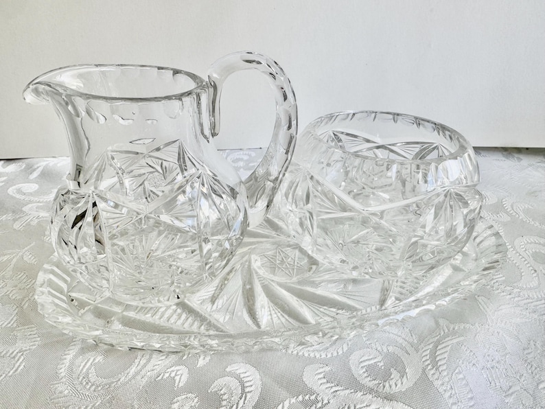 May include: A clear cut glass creamer pitcher and a matching sugar bowl set resting on an oval glass tray. Each piece features a detailed starburst and geometric faceted pattern with scalloped edges and a sturdy handle on the pitcher.