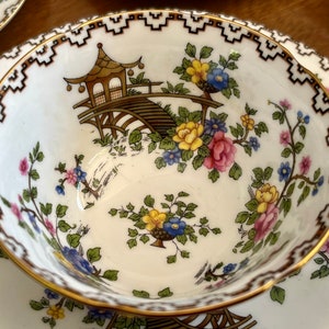K&ouml;nnte beinhalten: Eine wei&szlig;e Teetasse und Untertasse mit goldfarbenem Rand und einem komplizierten schwarzen und goldenen Randmuster. Die Tasse zeigt ein Blumenmuster mit rosa, gelben und blauen Blumen und einer braunen und goldenen Pagodenbr&uuml;cke.