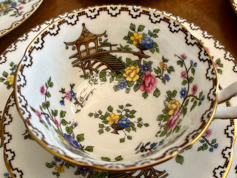 K&ouml;nnte beinhalten: Eine wei&szlig;e Teetasse und Untertasse mit goldfarbenem Rand und einem komplizierten schwarzen und goldenen Randmuster. Die Tasse zeigt ein Blumenmuster mit rosa, gelben und blauen Blumen und einer braunen und goldenen Pagodenbr&uuml;cke.