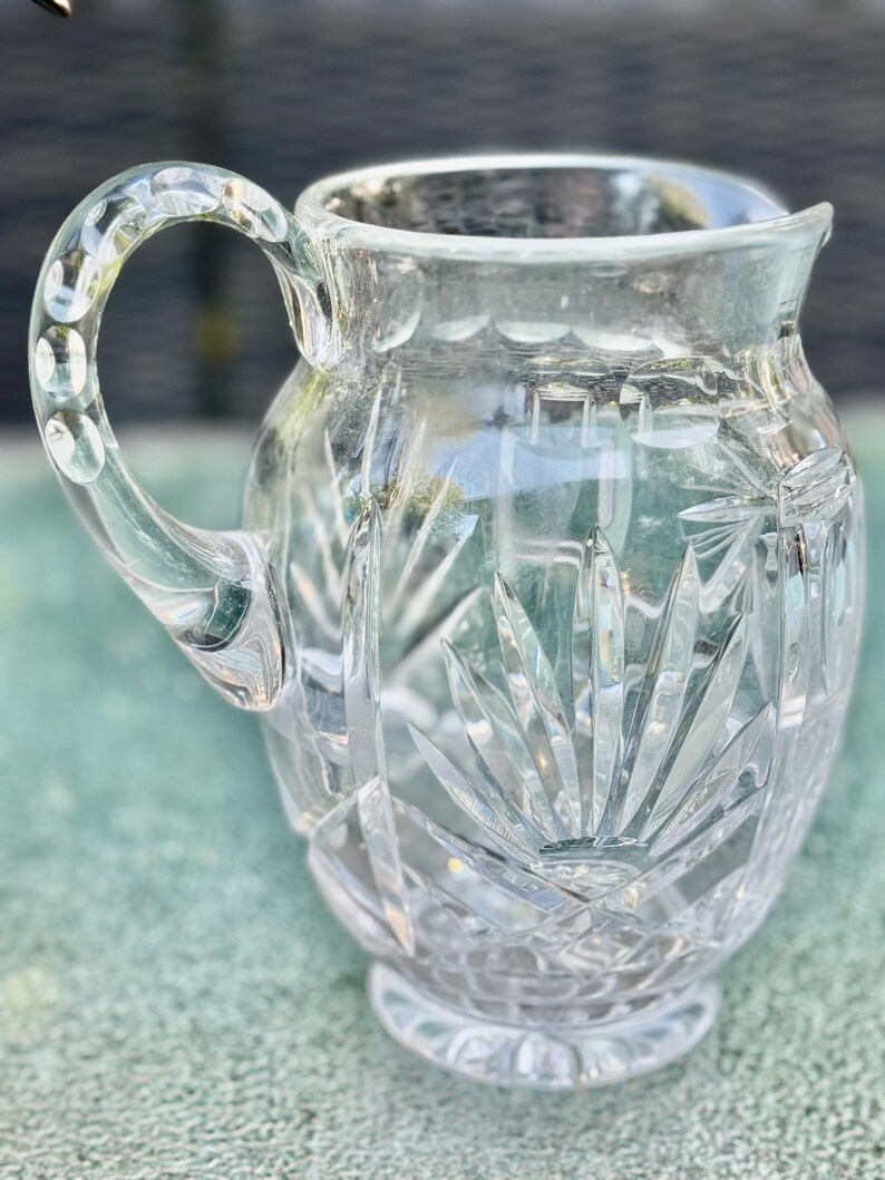 Puede incluir: Jarra de vidrio transparente con un asa decorativa y un dise&ntilde;o tallado. La jarra tiene una base ancha y una parte superior acampanada. El patr&oacute;n tallado presenta un dise&ntilde;o de estrella en la parte delantera y los lados. El asa tiene una serie de hendiduras redondeadas.