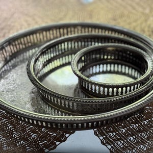 May include: A set of three nested round silver-toned metal trays with openwork gallery rails and beaded edges. The trays feature a reflective surface with an embossed floral pattern on the base of the largest tray and smooth centers.
