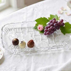 May include: A clear glass serving tray with a woven design, holding grapes, a pink rose, and three chocolates. A small floral plate with a chocolate is on the left. The scene is set on a white tablecloth.
