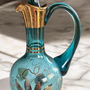 May include: A teal blue glass pitcher with a gold rim and gold leaf design. The pitcher has a round body and a curved handle.