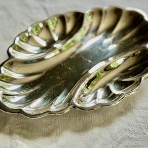 Pode incluir: Um prato de servir em metal prateado polido com formato oval e borda festonada semelhante a uma concha. A superf&iacute;cie &eacute; lisa e reflexiva, apresentando uma &aacute;rea central plana rodeada por reentr&acirc;ncias curvas e caneladas ao longo do per&iacute;metro.