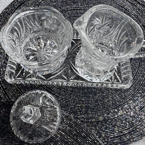 May include: A clear glass creamer and sugar bowl set with a matching rectangular tray. The pieces feature intricate starburst and fan-cut patterns. The sugar bowl includes a separate lid with a faceted knob handle, and the creamer has a handle.