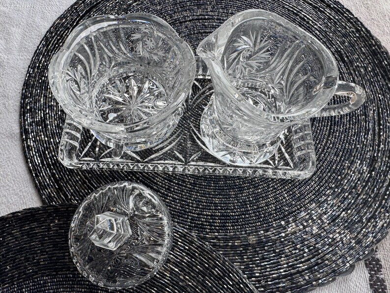 May include: A clear glass creamer and sugar bowl set with a matching rectangular tray. The pieces feature intricate starburst and fan-cut patterns. The sugar bowl includes a separate lid with a faceted knob handle, and the creamer has a handle.