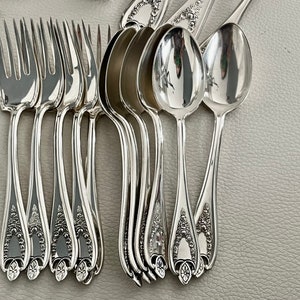 Puede incluir: Un conjunto de cubiertos antiguos de plata, que incluye tenedores y cucharas, dispuestos sobre una superficie gris claro. La cuberter&iacute;a tiene una superficie brillante y reflectante, con detalles florales ornamentados en los mangos. Los tenedores tienen cuatro p&uacute;as.