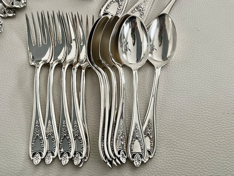 Puede incluir: Un conjunto de cubiertos antiguos de plata, que incluye tenedores y cucharas, dispuestos sobre una superficie gris claro. La cuberter&iacute;a tiene una superficie brillante y reflectante, con detalles florales ornamentados en los mangos. Los tenedores tienen cuatro p&uacute;as.