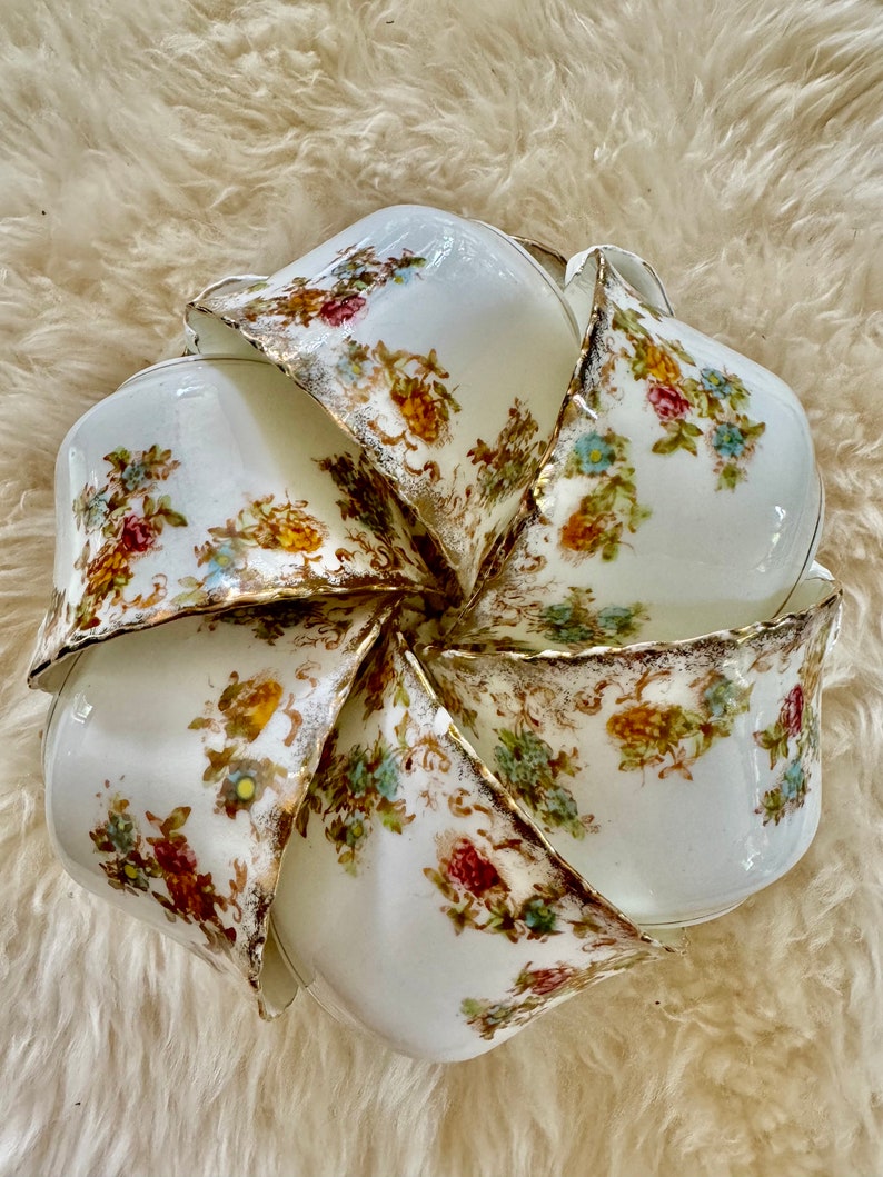 K&ouml;nnte beinhalten: Ein Set aus acht wei&szlig;en Porzellan-Teetassen mit floralem Muster und goldener Verzierung. Die Tassen sind in einem Kreis angeordnet, wobei die R&auml;nder der Tassen nach au&szlig;en zeigen.