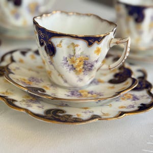 May include: A vintage teacup and saucer set. The white porcelain features a navy blue rim and gold detailing. The floral design includes yellow and purple flowers. The saucer has a scalloped edge. A classic tea set for serving tea.
