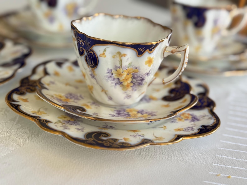 May include: A vintage teacup and saucer set. The white porcelain features a navy blue rim and gold detailing. The floral design includes yellow and purple flowers. The saucer has a scalloped edge. A classic tea set for serving tea.