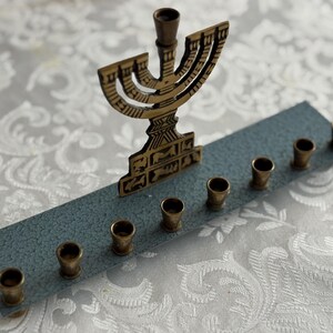 May include: A brass menorah with nine candle holders, set on a blue-grey base. The menorah is a traditional Jewish candelabrum, used during the holiday of Hanukkah. The background is a white, patterned tablecloth.