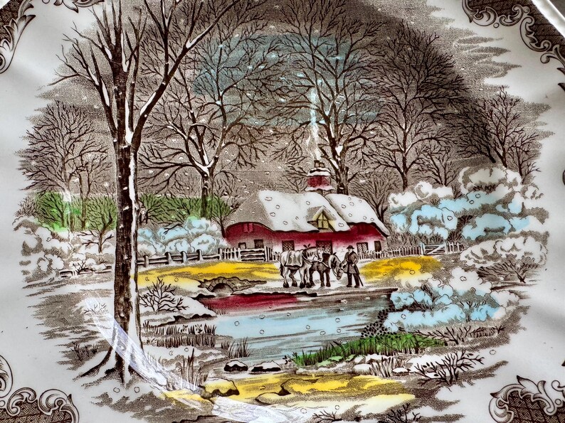 May include: A vintage ceramic plate with a winter scene. The plate features a snow-covered farmhouse with a red roof, trees, and a frozen pond. The scene is painted in brown, blue, and white tones.