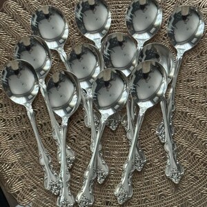 May include: A collection of twelve silver spoons with ornate, floral-patterned handles. The spoons have a reflective, polished surface, and are arranged on a textured, gold-colored surface. The spoons are designed for serving.