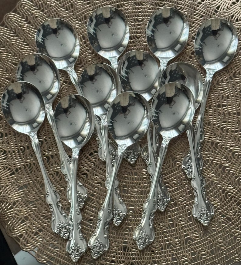 May include: A collection of twelve silver spoons with ornate, floral-patterned handles. The spoons have a reflective, polished surface, and are arranged on a textured, gold-colored surface. The spoons are designed for serving.