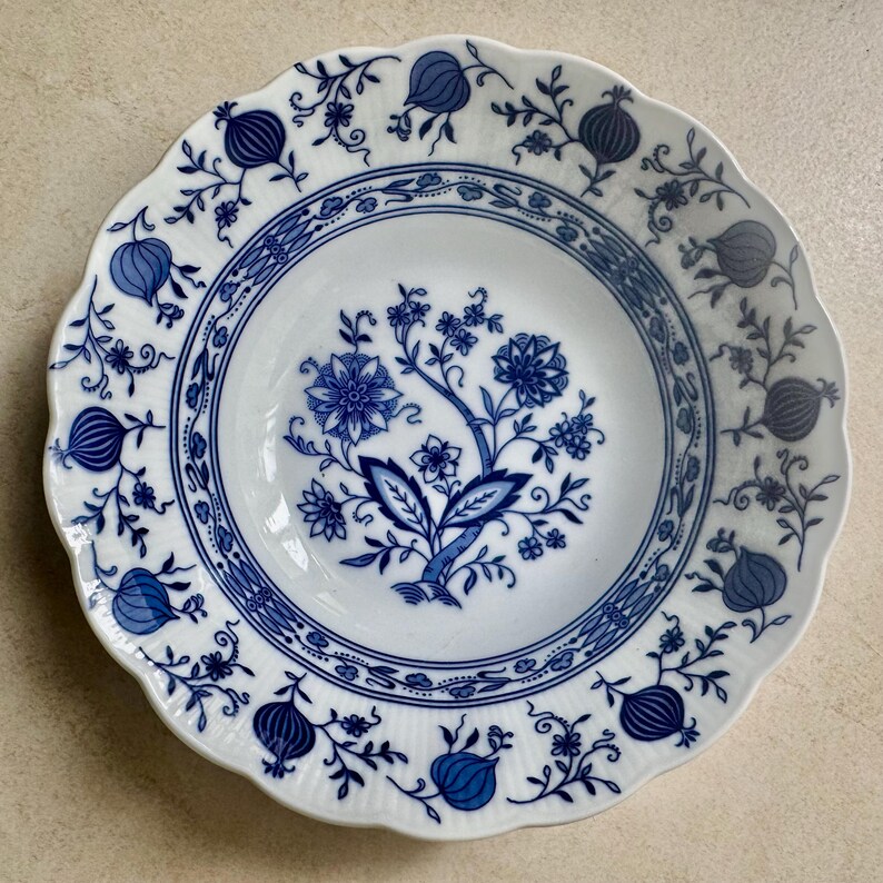 May include: A white ceramic bowl with a scalloped edge featuring a blue floral pattern. The center displays a detailed botanical motif, while the rim is decorated with repeating pomegranate-like shapes and delicate vine-like floral accents.