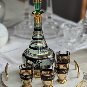 May include: A set of four clear glass shot glasses with gold and black stripes and a matching green and gold glass decanter with a gold stopper. The glasses and decanter are on a white marble tray with a gold rim.