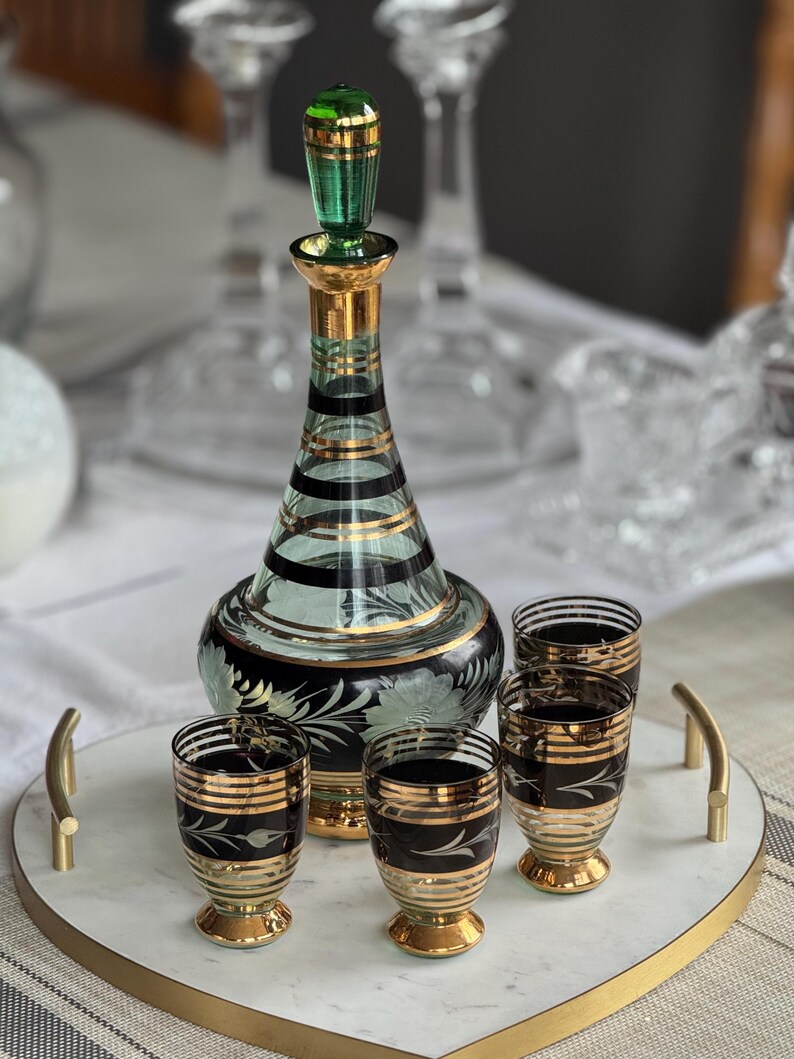 May include: A set of four clear glass shot glasses with gold and black stripes and a matching green and gold glass decanter with a gold stopper. The glasses and decanter are on a white marble tray with a gold rim.
