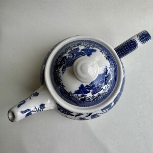 K&ouml;nnte beinhalten: Eine wei&szlig;e Teekanne mit einem blauen und wei&szlig;en Weidenmuster. Die Teekanne hat einen Deckel und einen Ausguss. Der Griff befindet sich an der Seite der Teekanne.