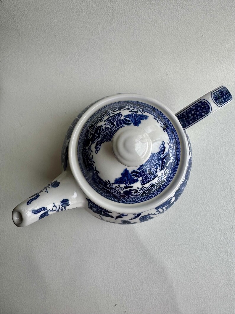 K&ouml;nnte beinhalten: Eine wei&szlig;e Teekanne mit einem blauen und wei&szlig;en Weidenmuster. Die Teekanne hat einen Deckel und einen Ausguss. Der Griff befindet sich an der Seite der Teekanne.