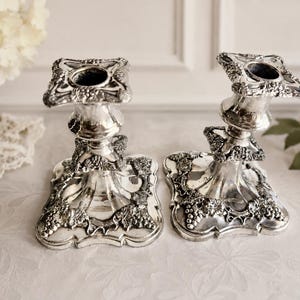 May include: A pair of silver-toned metal candle holders featuring a square base and top, both decorated with embossed grape and vine leaf motifs. The candle holders have a polished, reflective finish and a flared design that tapers toward the center.