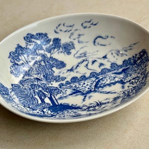 May include: Oval, white ceramic serving dish with a blue landscape design. The design features trees, a bridge, and mountains. The dish is approximately 10 inches long and 7 inches wide.