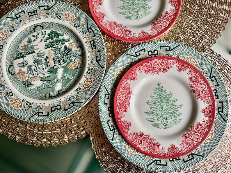 May include: A collection of decorative plates. One plate features a green and white Asian-inspired landscape design. Other plates have a red floral border and a green Christmas tree design. The plates are arranged on a gold-colored placemat.
