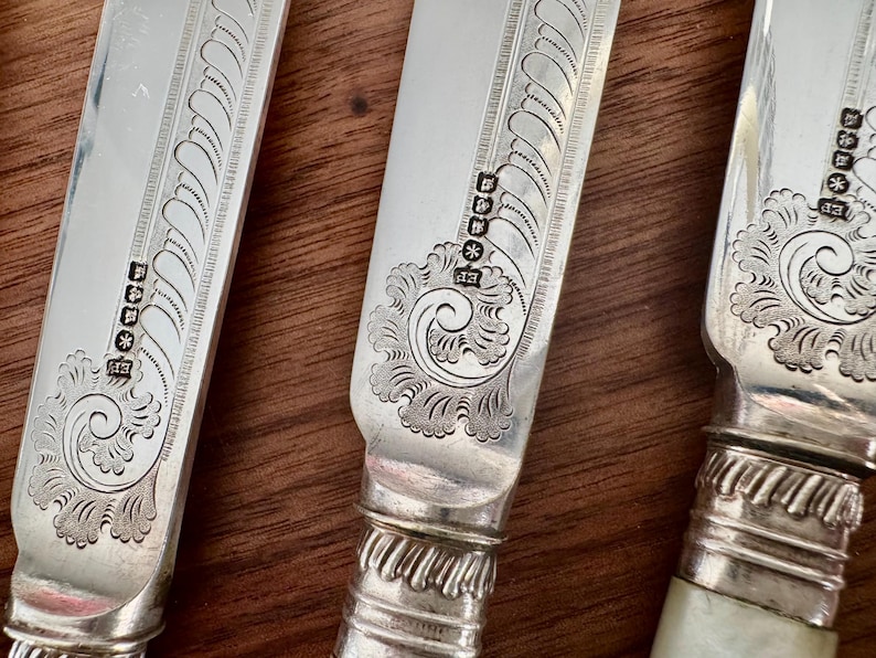 Pu&ograve; includere: Primo piano di tre coltelli da tavola in argento con disegni decorativi e punzoni. I coltelli hanno un manico in argento e una lama con un motivo decorativo.