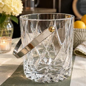 May include: A clear glass ice bucket featuring a textured, crisscross geometric pattern on the exterior. The bucket includes a polished silver-tone metal swing handle attached with gold-tone circular hardware on both sides of the cylindrical vessel.