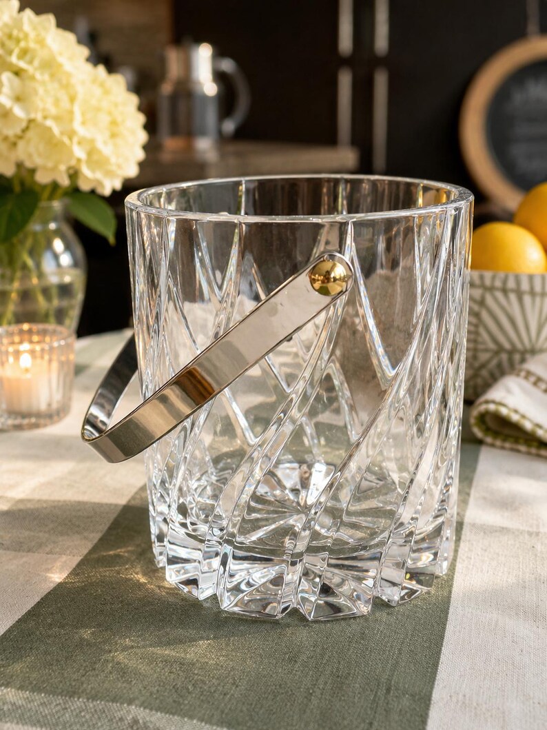 May include: A clear glass ice bucket featuring a textured, crisscross geometric pattern on the exterior. The bucket includes a polished silver-tone metal swing handle attached with gold-tone circular hardware on both sides of the cylindrical vessel.