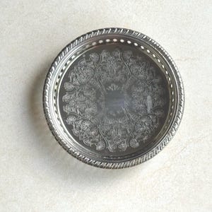 May include: A round silver-toned metal tray featuring an intricate engraved floral pattern across the center. The rim has a raised, textured border with a series of small, evenly spaced cut-out holes circling the entire edge of the tray.