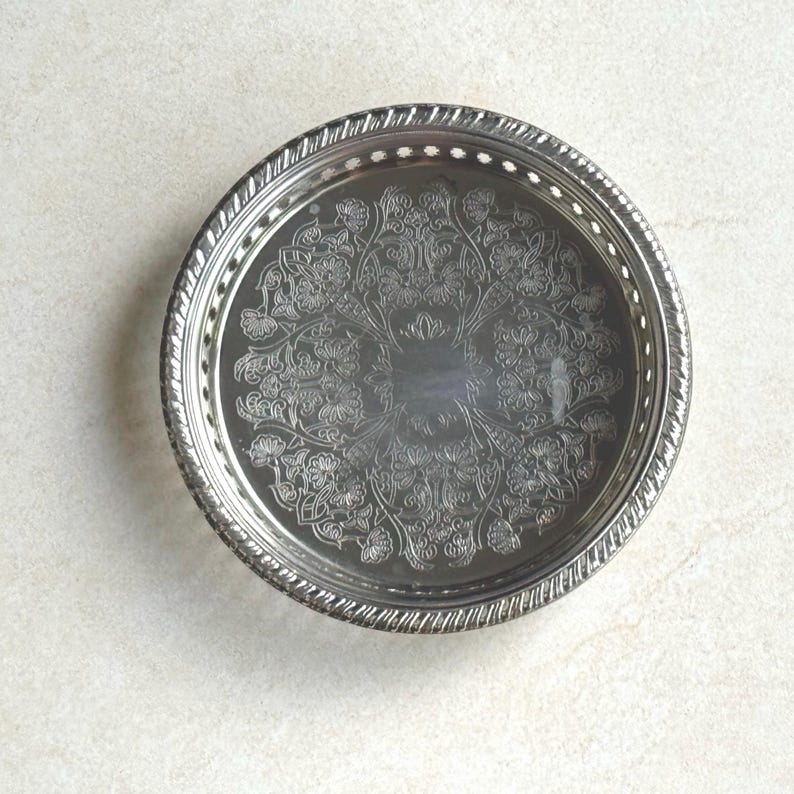 May include: A round silver-toned metal tray featuring an intricate engraved floral pattern across the center. The rim has a raised, textured border with a series of small, evenly spaced cut-out holes circling the entire edge of the tray.
