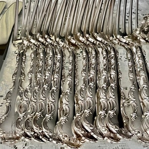 May include: An array of ornate silver forks and knives with decorative handles. The silverware is arranged on a white lace tablecloth, reflecting light. The forks and knives have a detailed, floral design.