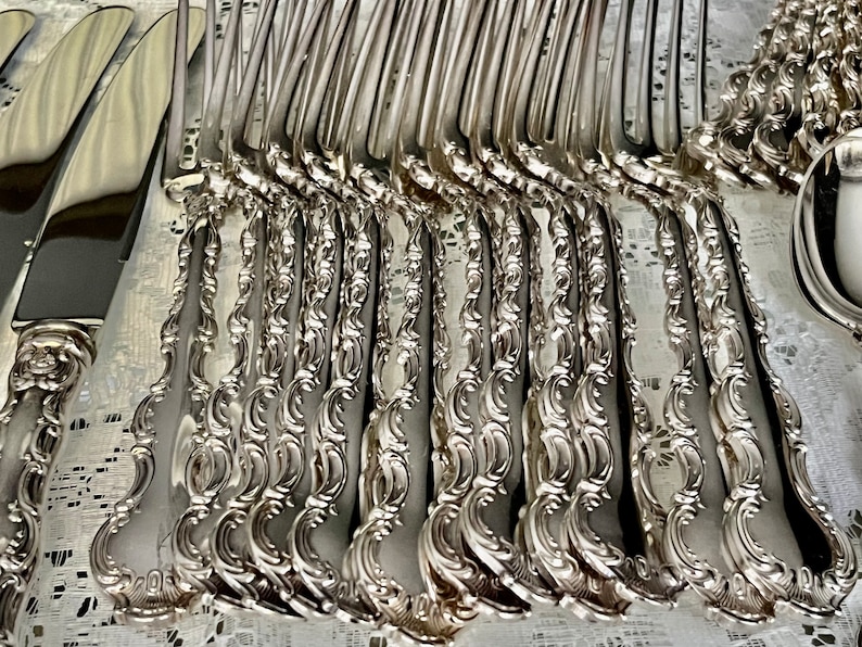 May include: An array of ornate silver forks and knives with decorative handles. The silverware is arranged on a white lace tablecloth, reflecting light. The forks and knives have a detailed, floral design.