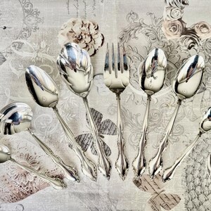 May include: An assortment of antique silver-plated serving utensils, including spoons and a fork, arranged on a patterned surface. The spoons have ornate, detailed handles and reflective bowls. The fork has four tines. The overall aesthetic is vintage and elegant.