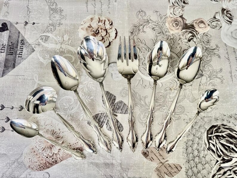 May include: An assortment of antique silver-plated serving utensils, including spoons and a fork, arranged on a patterned surface. The spoons have ornate, detailed handles and reflective bowls. The fork has four tines. The overall aesthetic is vintage and elegant.