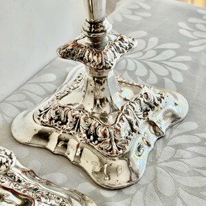 May include: A pair of silver-toned metal candle holders featuring a square base with ornate, raised scrollwork and floral relief detailing. The reflective surface shows a polished finish with a tapered stem rising from the center of each base.