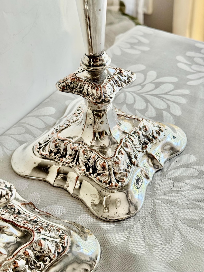 May include: A pair of silver-toned metal candle holders featuring a square base with ornate, raised scrollwork and floral relief detailing. The reflective surface shows a polished finish with a tapered stem rising from the center of each base.