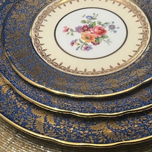 May include: A stack of vintage china plates with a blue and gold floral pattern. The plates have a scalloped edge and a gold rim. The center plate features a cream-colored background with a floral bouquet.