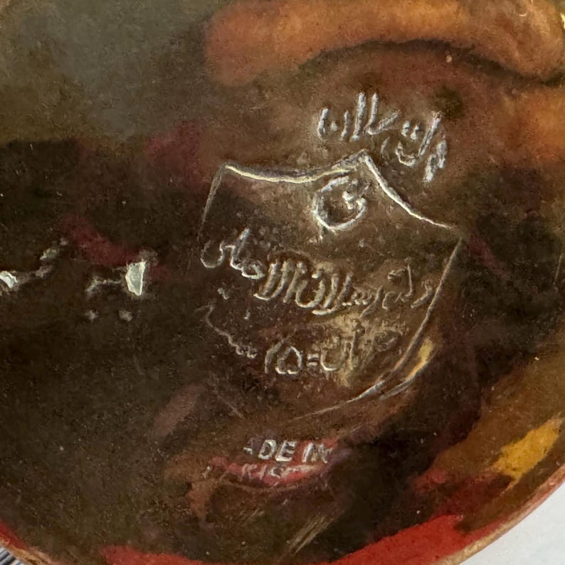 Może przedstawiać: Zbliżenie metalowego przedmiotu, prawdopodobnie mosiężnego, z wytłoczonym arabskim pismem i emblematem tarczy. Widoczny jest napis "MADE IN", wskazujący na pochodzenie przedmiotu. Obiekt ma ciepły, postarzany wygląd.